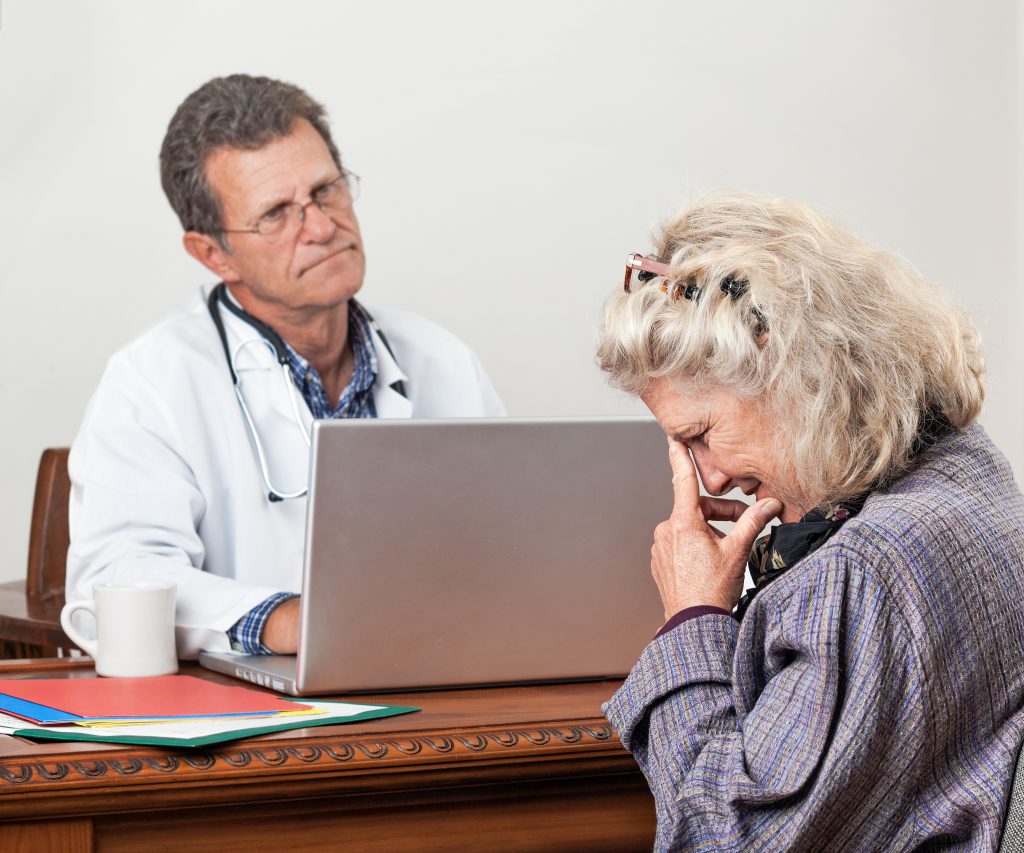 Pretty mature woman consults with her doctor in his office. Focus is on the woman’s face. She looks worried and tearful. The doctor looks bored and impatient.
