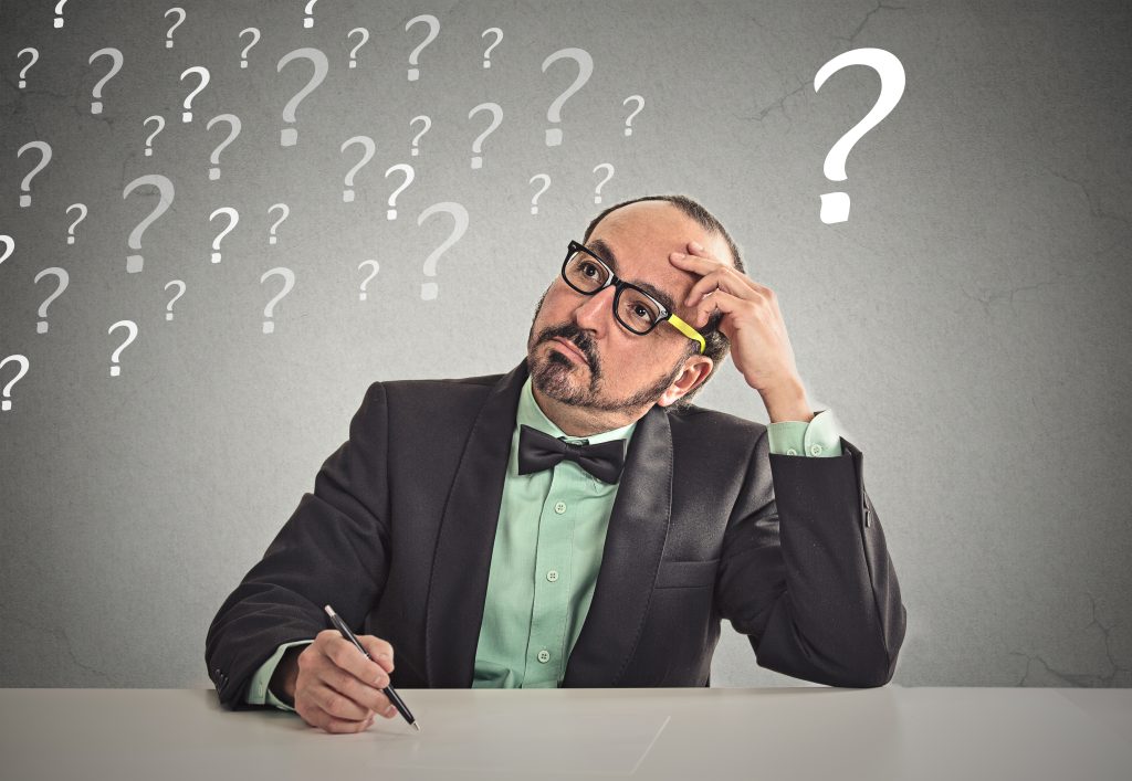 Confused puzzled business man sitting at table scratching his head thinking has many questions isolated office grey wall background. Human face expression emotion feeling body language perception