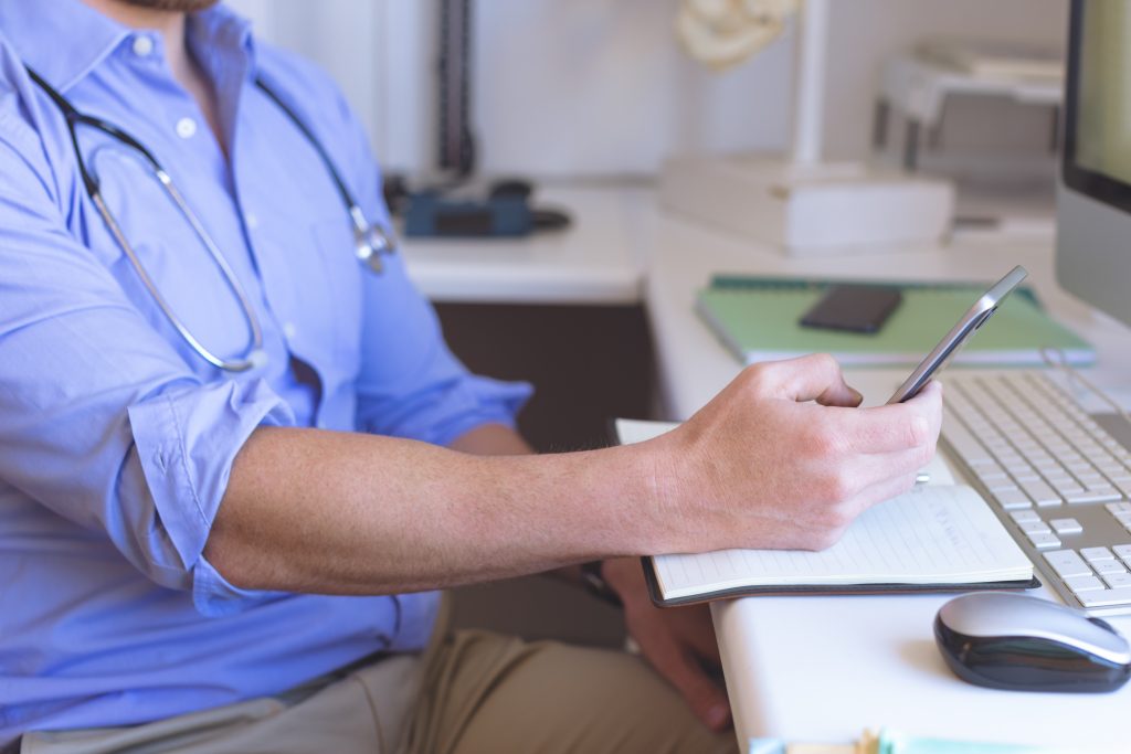 Mid-section of confident Caucasian male doctor using mobile phone in clinic against his desk in background